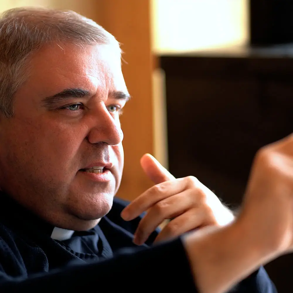 Ksiądz w koloratce podczas rozmowy o poznaniu Boga w liturgii; gestykuluje, podkreślając zmysłowy wymiar liturgii.